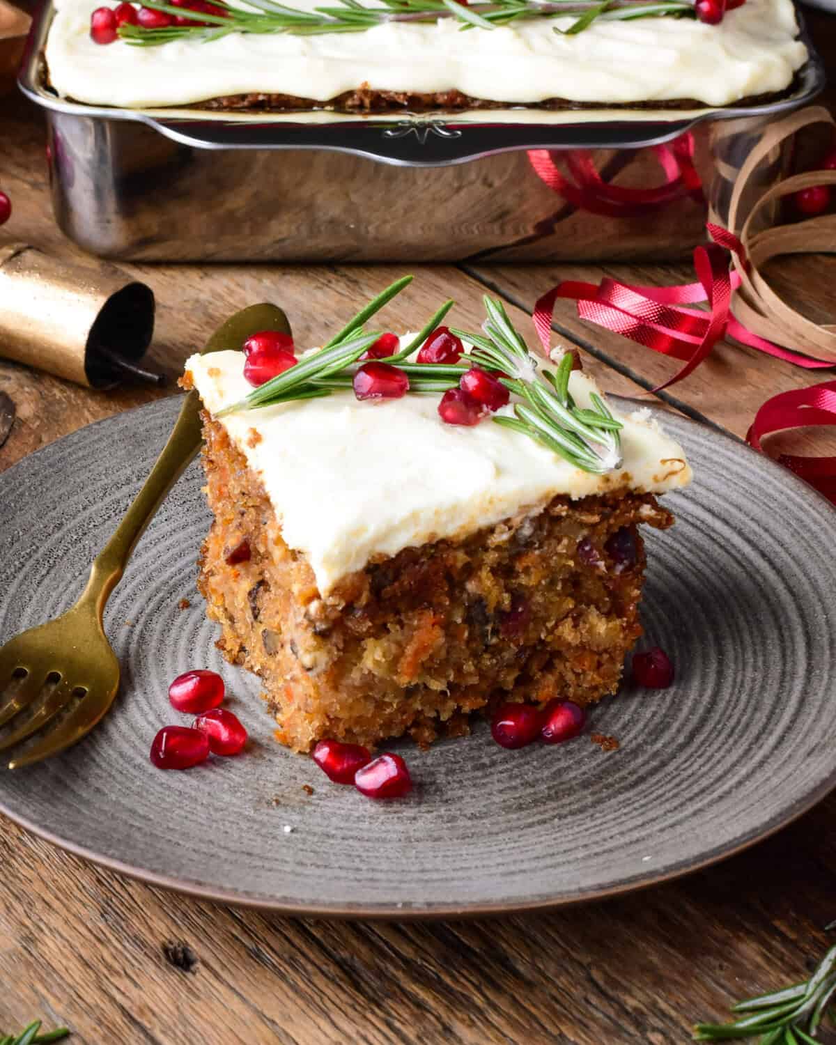 A corner slice of a Christmas carrot cake with cream cheese icing. Decorated with pomegranate arils and rosemary.
