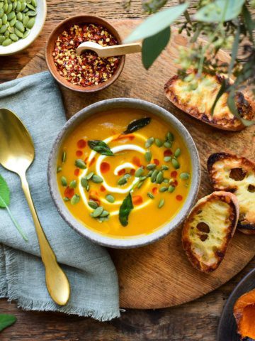 A bowl of Honeynut Squash Soup topped with a swirl of Greek yogurt, sprinkled with pepitas, chili oil and fried sage. Extra topping and three slices of baguette toast are surrounding the bowl.