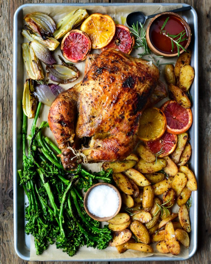 A rotisserie chicken on a platter with grilled vegetables and orange slices surrounding it.