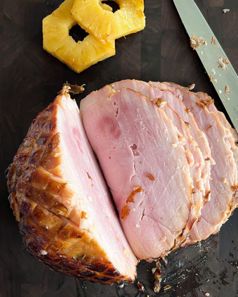 A baked ham with pineapple slices. Half of the ham is sliced.  