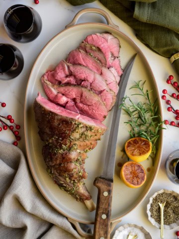 A medium rare Roasted Leg of Lamb with Lemon Vinaigrette half half-sliced on a platter with a knife.
