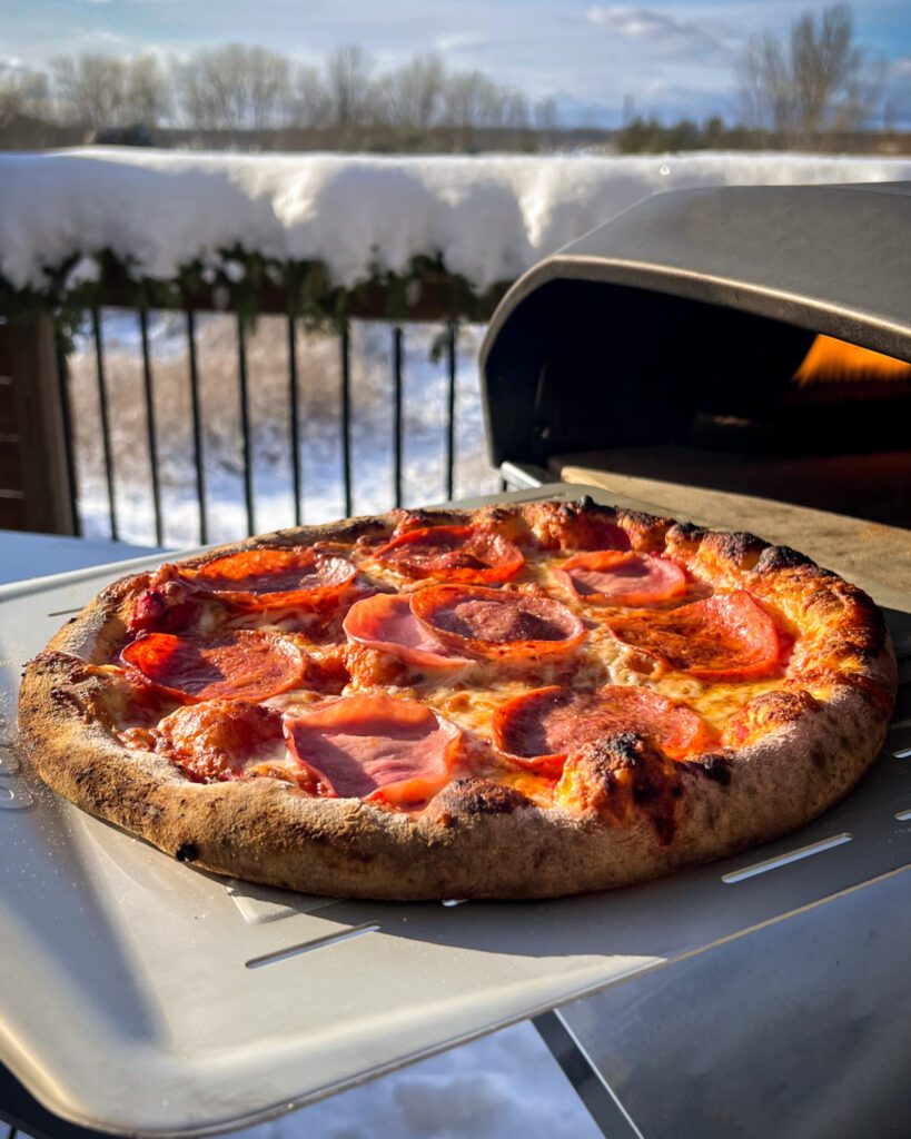 A 3 meat pizza coming out of a flaming Ooni pizza oven.