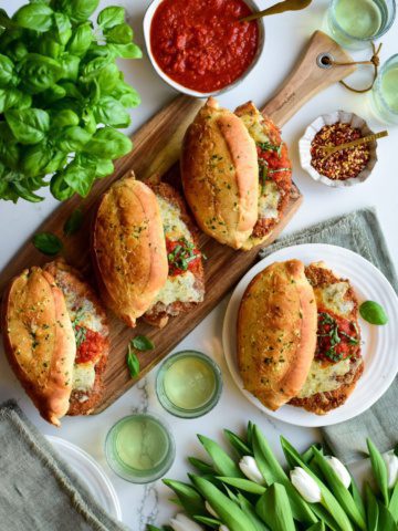 Three crispy chicken sandwiches that look like chicken parmesan sandwiches on a tray, and one is on a plate. Served with extra sauce, basil, and drinks.