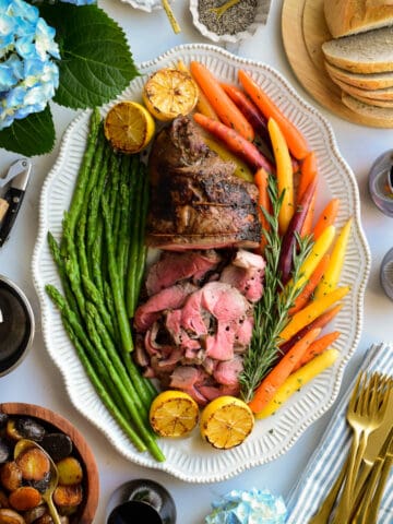 An oval platter with a half sliced rotisserie boneless leg of lamb served with heirloom carrots, asparagus and roasted lemons.