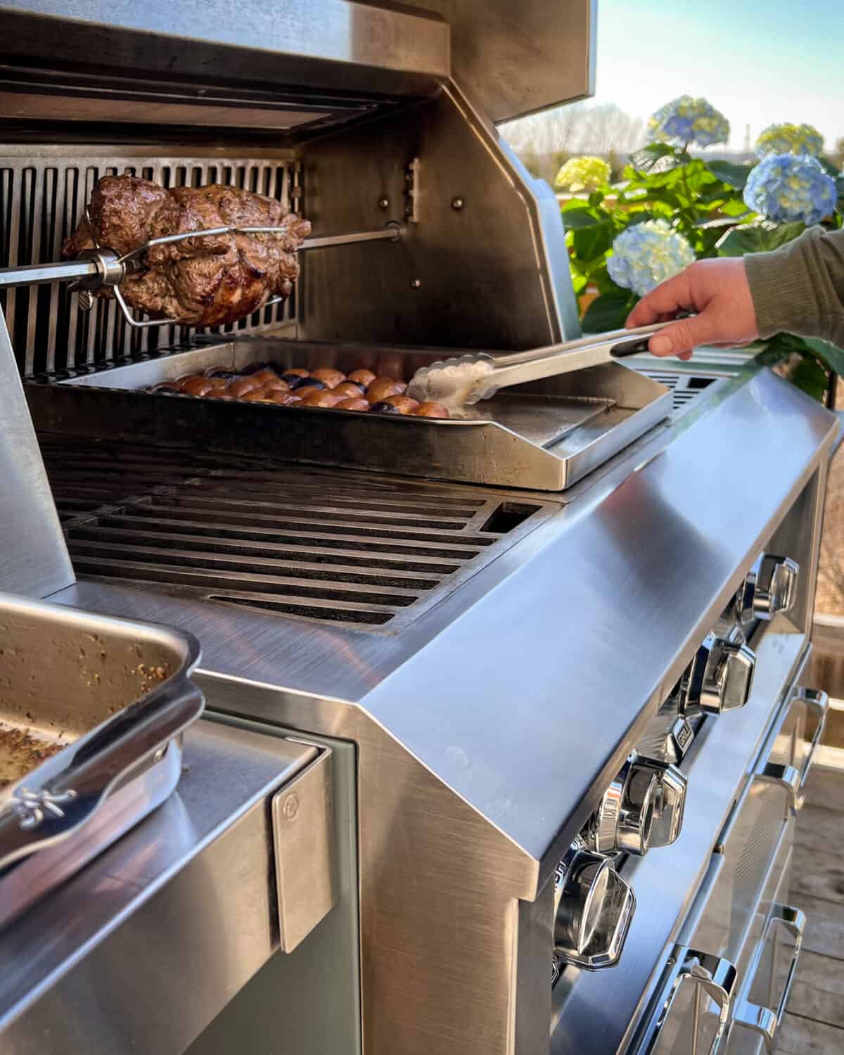 Side shot of a boneless leg of lamb on the rotisserie with a griddle of potatoes below being tossed.