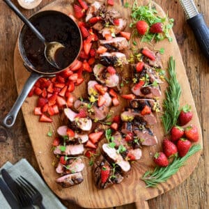 Sliced Sous-vide Pork Tenderloin with Strawberry-Balsamic Sauce on a board with a pot of sauce and fresh strawberries.
