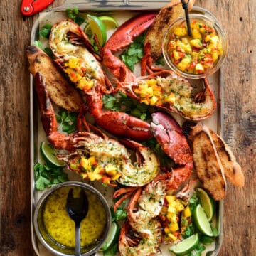 Grilled half lobsters topped with a Mango-Jalapeño Salsa on a tray with a pot of garlic-chive butter.