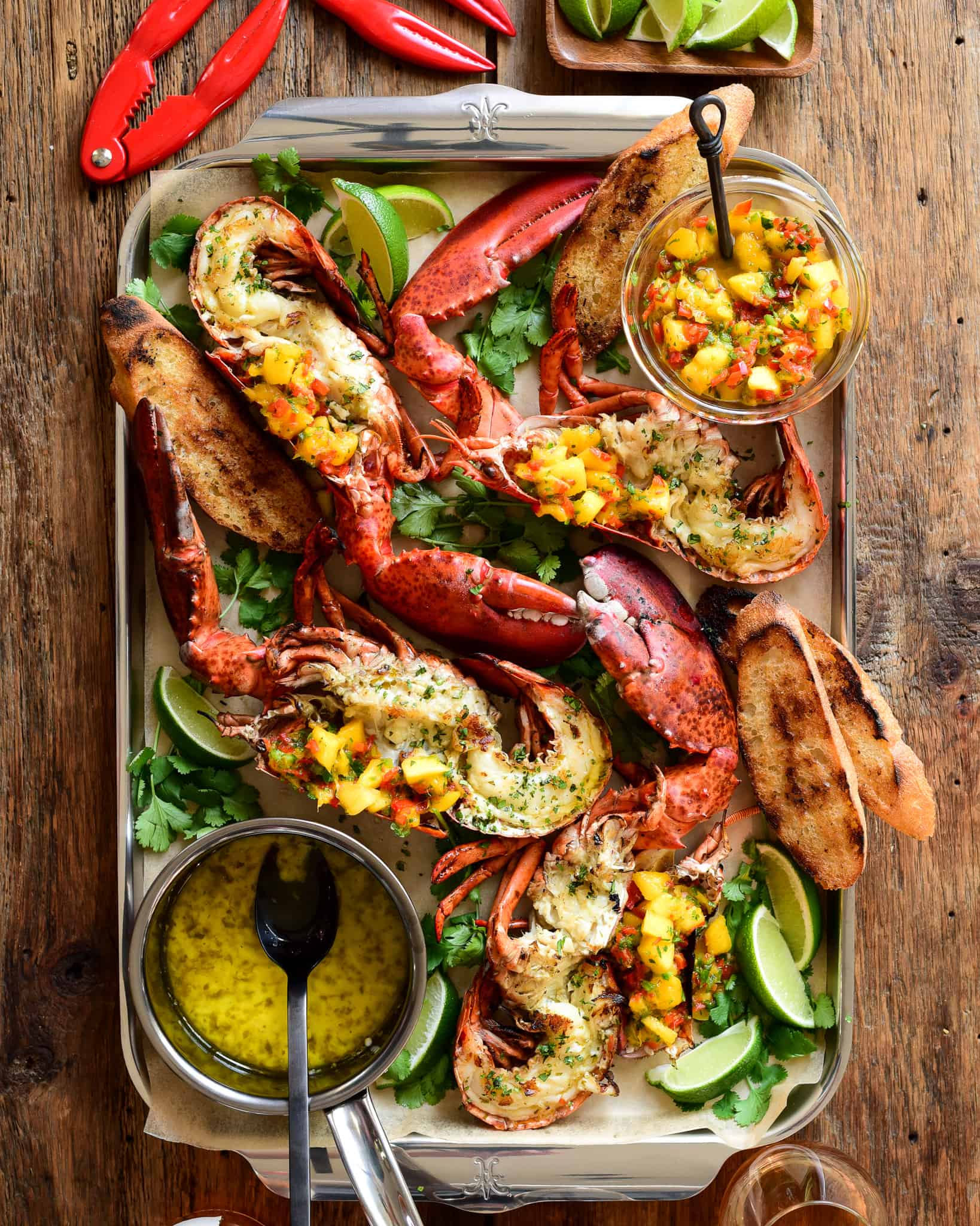 Grilled half lobsters topped with a Mango-Jalapeño Salsa on a tray with a pot of garlic-chive butter. 
