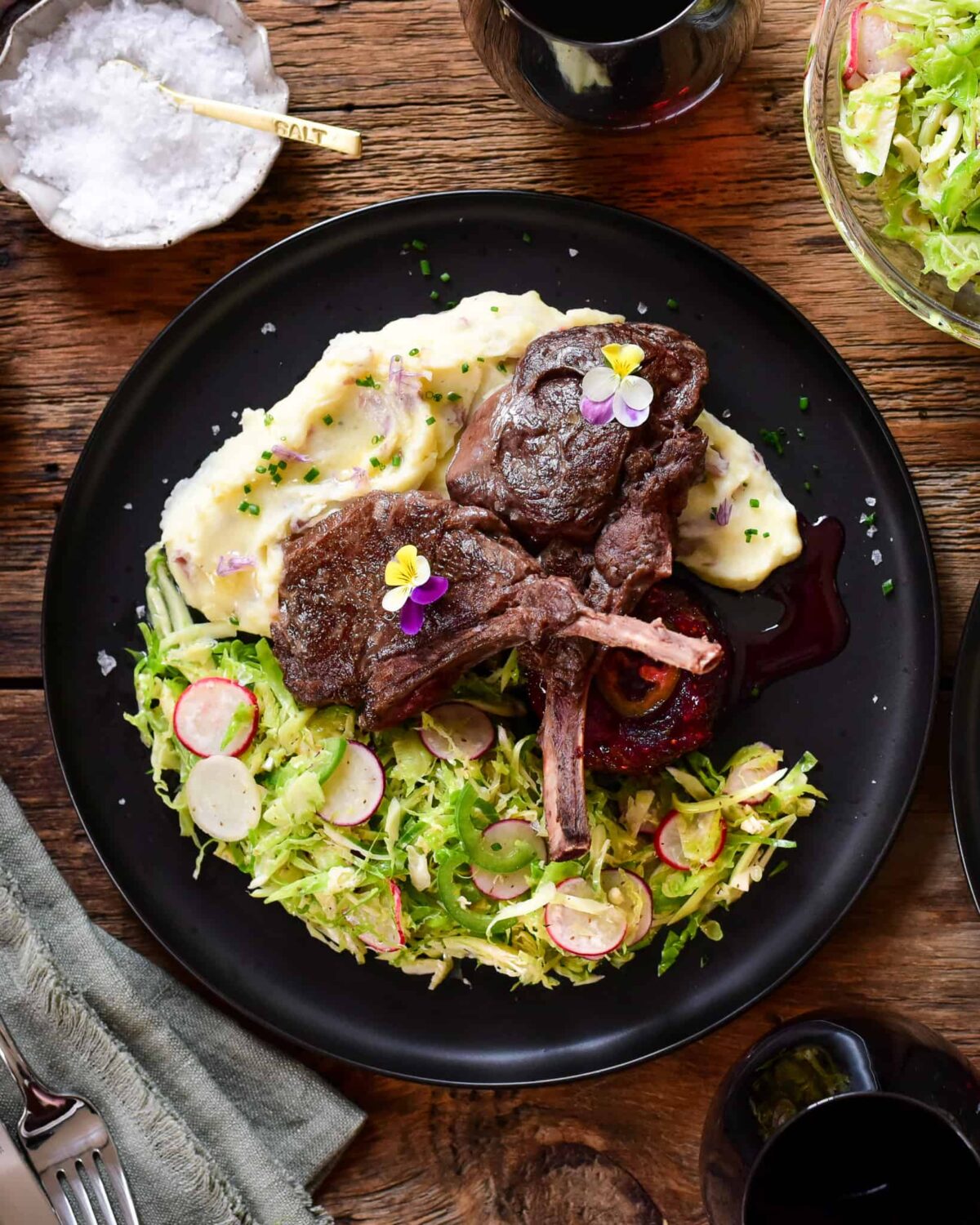 Two venison chops are leaning over mashed potatoes, a brussels sprout slaw alongside and Jalapeño Sauce on a black plate.