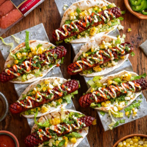 Six crosshatched beef smokies in taco holders dressed with Elote-style corn ingredients.