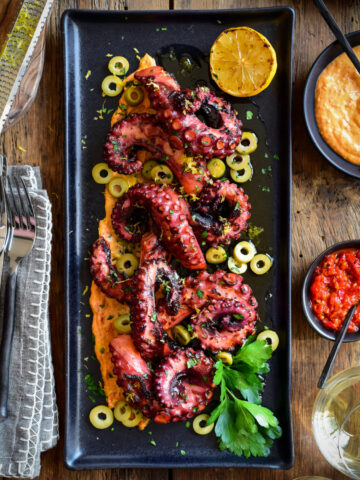 A black rectangular plate with grilled and very tender sous-vide octopus tentacles on a bed of spicy red pepper hummus.