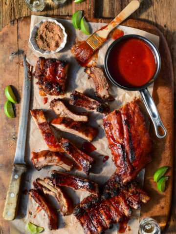 A cutting board with sauced baby back ribs and a pot of tequila BBQ sauce. A large knife has sliced most of the ribs.