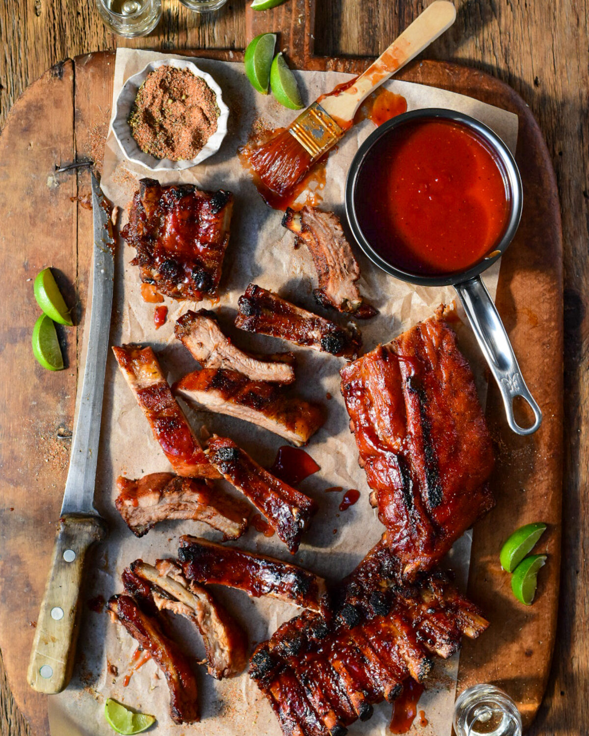 A cutting board with sauced baby back ribs and a pot of tequila BBQ sauce. A large knife has sliced most of the ribs. 