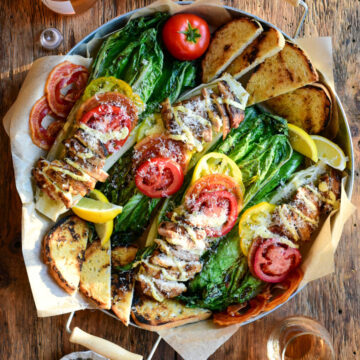 Four grilled romaine halves are topped with tomatoes, pancetta and chicken thighs and drizzled with a Caesar dressing, Parmesan and sourdough toasts in a round serving tray.