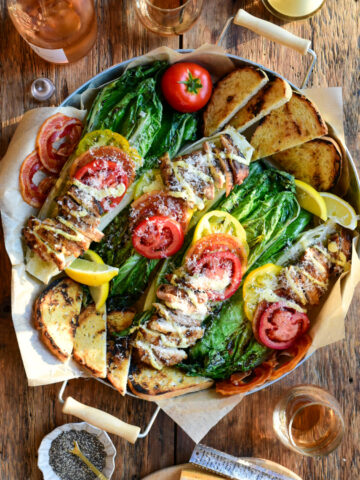Four grilled romaine halves are topped with tomatoes, pancetta and chicken thighs and drizzled with a Caesar dressing, Parmesan and sourdough toasts in a round serving tray.