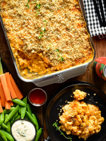 A tray of Buffalo Cauliflower Mac and Cheese with a portion on a plate. Served with crudités and blue cheese dip.