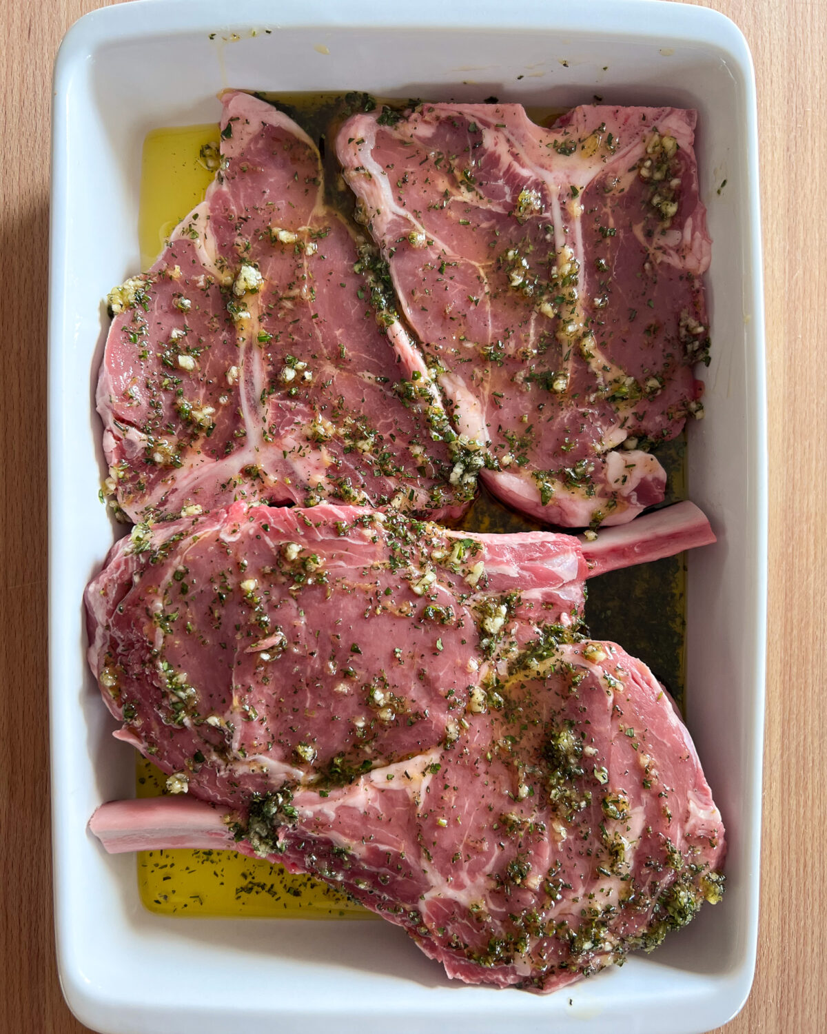 Four veal chops marinating in olive oil, chopped rosemary, chopped thyme, lemon zest, and garlic.