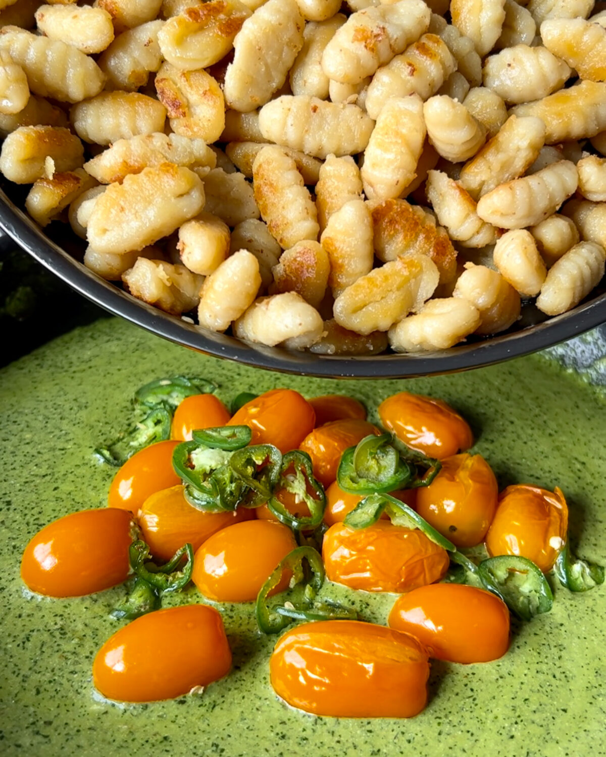 Adding in the browned gnocchi into a skillet with blistered tomatoes, jalapenos and pesto sauce.