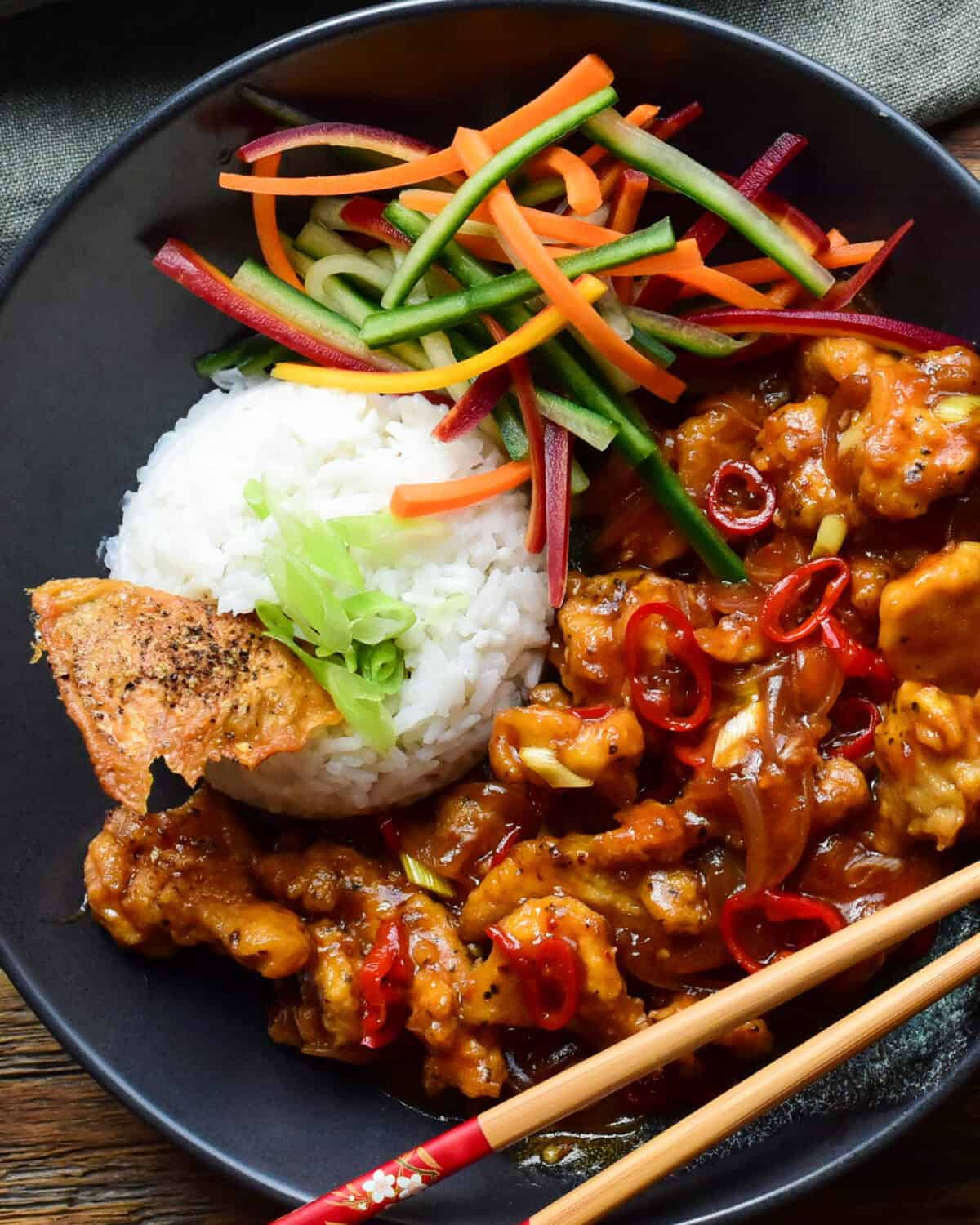 A close up of a bowl of Chili Chicken with Coconut Ginger Rice served with a carrot and cucumber salad and crispy chicken skins.