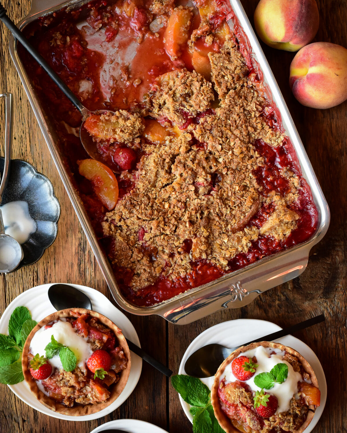 A baking tray with a baked Peach Strawberry Crisp with two bowls served with ice cream and mint.