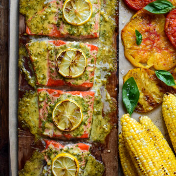 Cedar Plank Sockeye Salmon portioned in four with sauce and lemon slices. Served with grilled corn and tomatoes.
