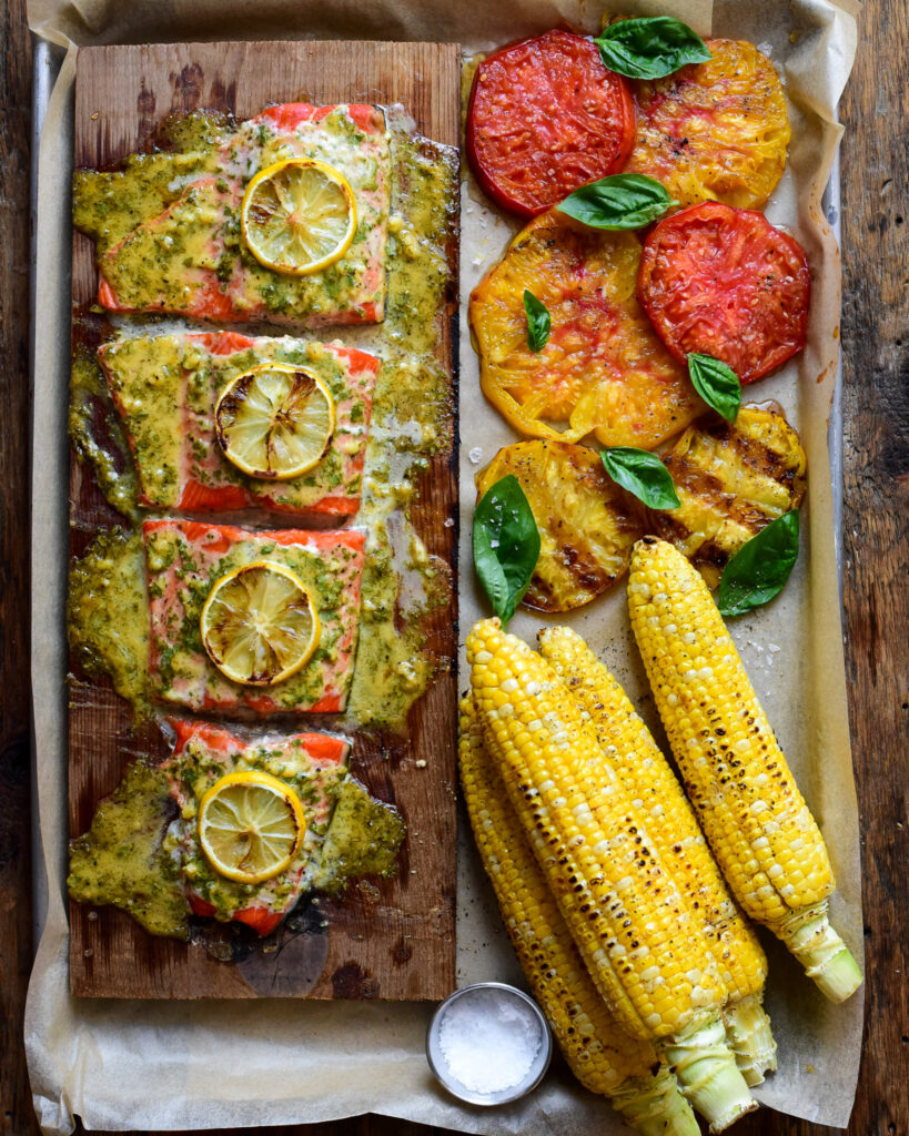 A tray with four pieces of cedar planked salmon served with grilled tomatoes and corn on the cob.