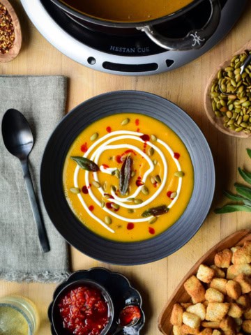 A bowl of Butternut Squash and Ginger Soup garnished with Greek yogurt, pepitas, chili oil, fried sage. Surrounded by toppings and croutons.