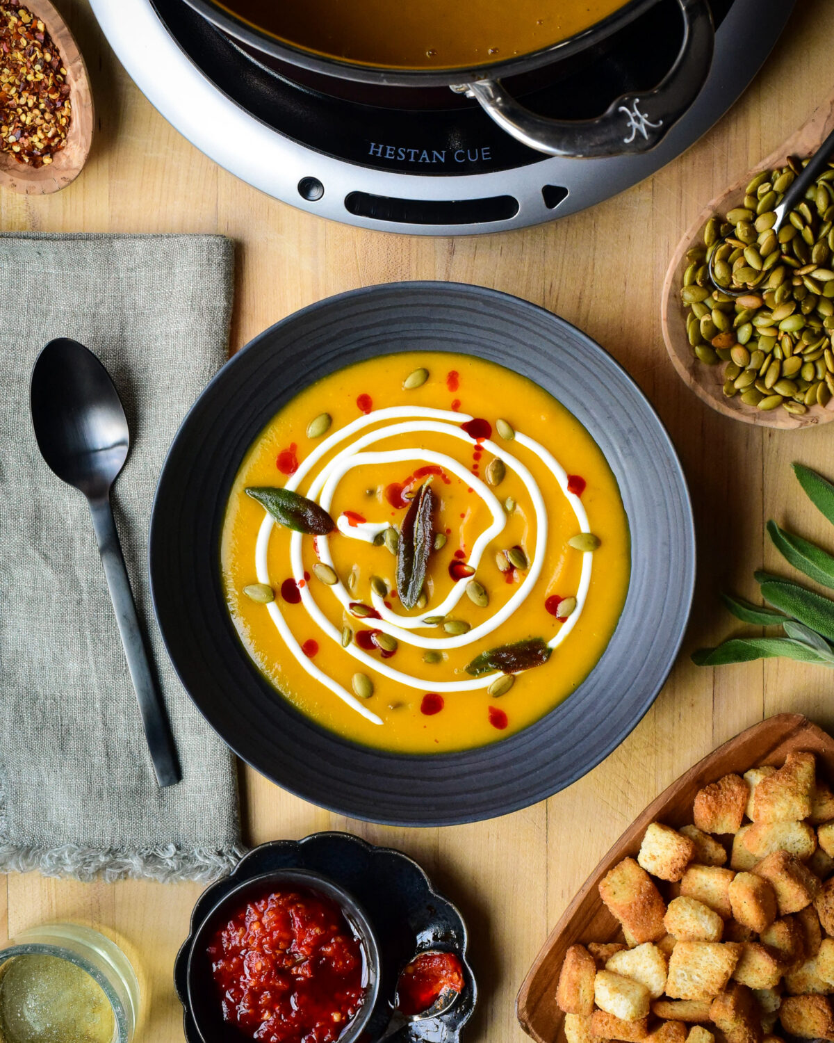 A bowl of Butternut Squash and Ginger Soup garnished with Greek yogurt, pepitas, chili oil, fried sage. Surrounded by toppings an croutons.