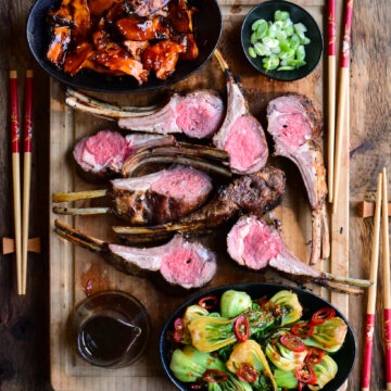 A serving board with two Grilled Frenched Rack of Lamb, cut open and served with Grilled Gochujang Mushrooms and Garlic-Ginger Bok Choy.
