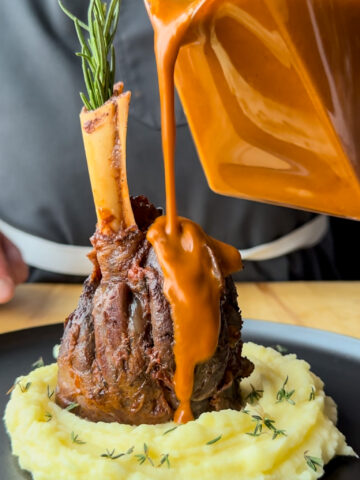 A braised lamb shank placed on a mound of mashed potatoes with sauce poured over it.