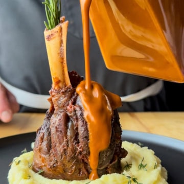 A braised lamb shank placed on a mound of mashed potatoes with sauce poured over it.