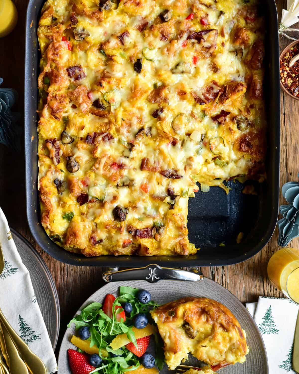 A roasting pan with a make ahead croissant breakfast casserole, with a piece taken out and served with fruit.