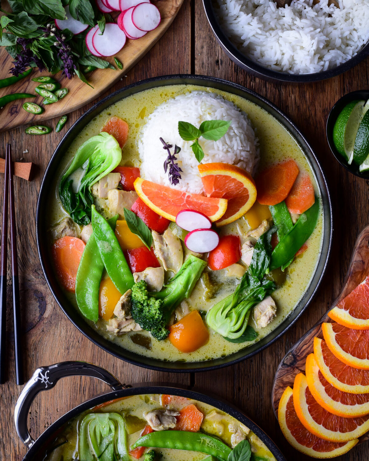 A colourful and vibrant plate of Thai Green Curry Chicken & Vegetables. Served with jasmine rice, Thai basil, orange slices and green chilies.