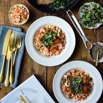 Two plated dishes of Ora King Salmon with Spinach & Chow-Chow. A Thomas Keller skillet and The French Laundry cook book are also on the table.