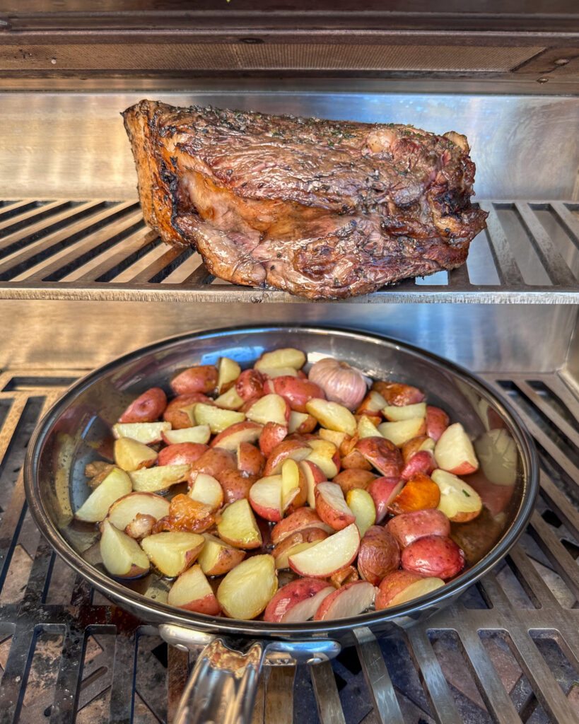 A large ribeye on the upper rack of the grill with a skillet of potatoes underneath to catch the drippings.