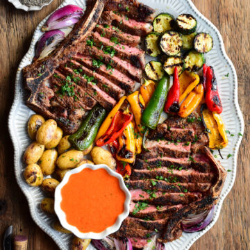 Two sliced Spanish Style Steaks with assorted grilled vegetables and a bowl of Spanish steak sauce.