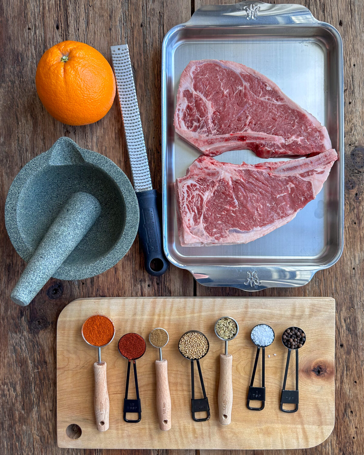 Ingredients for a steak rub: several spices, two steaks, an orange, a mortar and pestle and a micro plane.