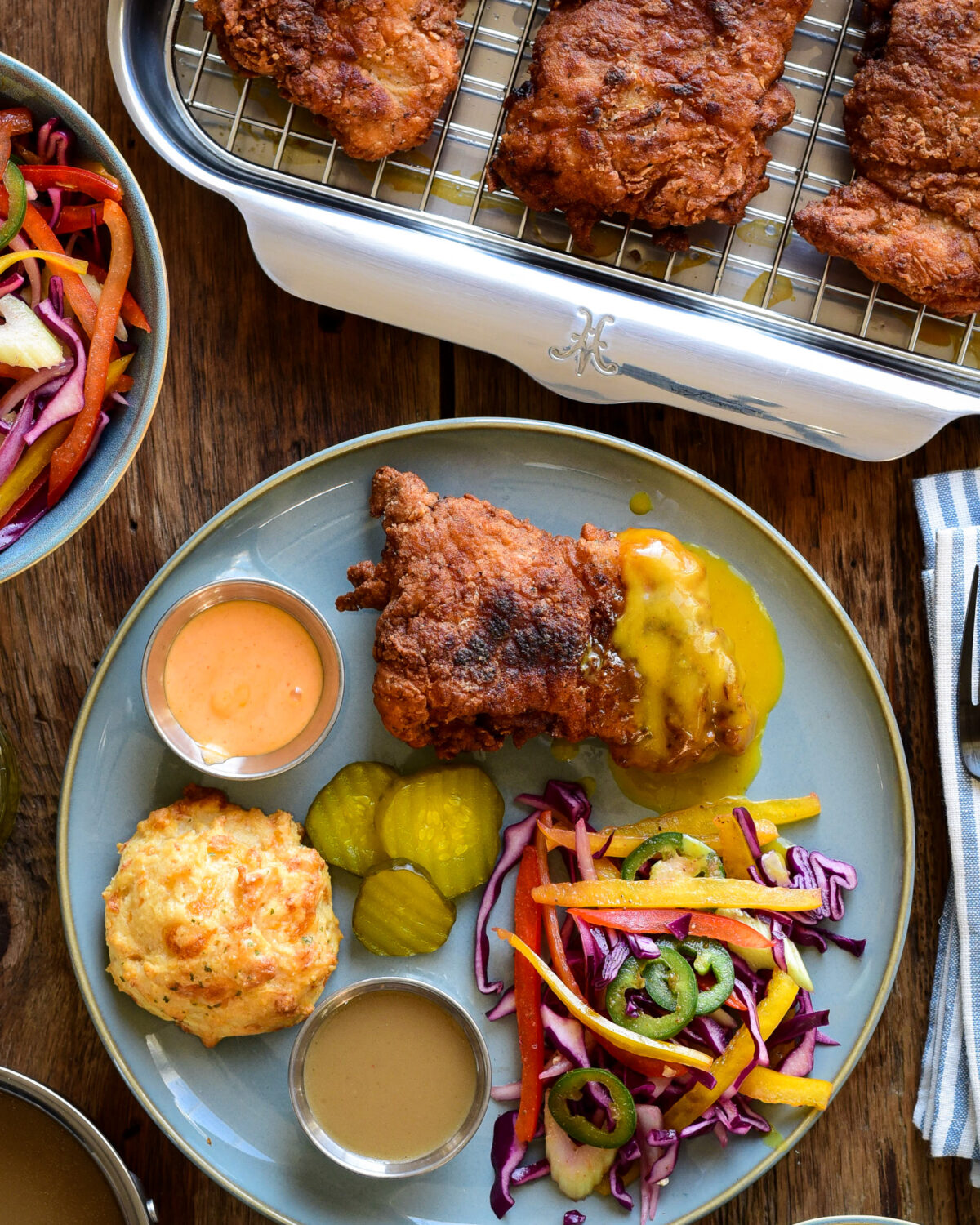 A plate with a piece of spicy buttermilk fried chicken served with a psychedelic slaw, cheddar biscuits and three dips.