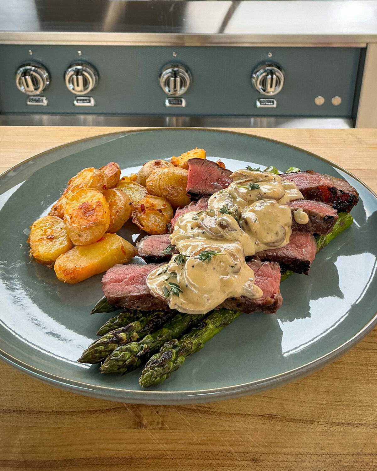 A plate with grilled steak slices overtop asparagus with a mushroom sauce and bomba potatoes.