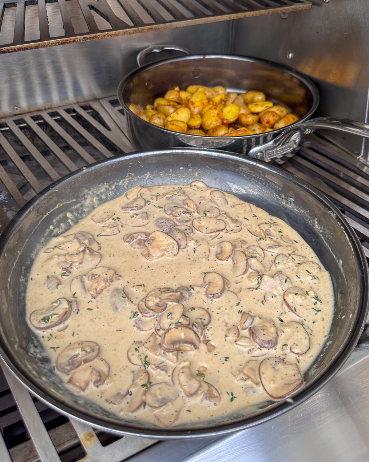 Two skillets on a grill, one with a mascarpone mushroom sauce and the other with bomba potatoes.