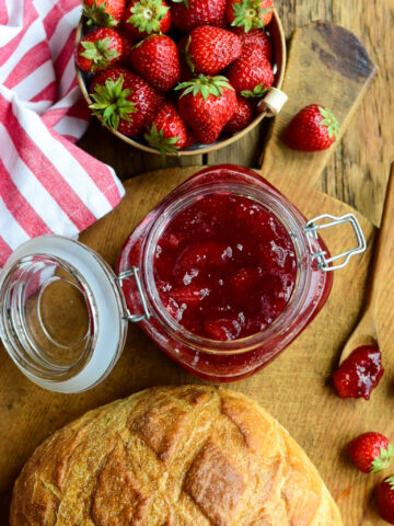 A large mason jar filled with Strawberry-Ginger Jam on a board with a sourdough loaf and fresh strawberries.