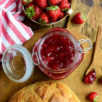 A large mason jar filled with Strawberry-Ginger Jam on a board with a sourdough loaf and fresh strawberries.