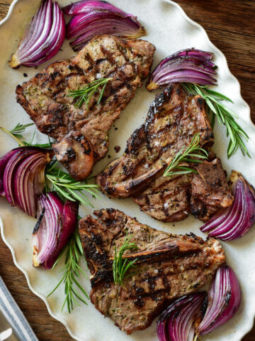 A platter with three grilled veal t-bone chops with grilled red onions and rosemary.
