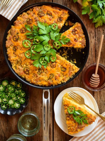 A skillet with a grilled Jalapeño Cheddar Cornbread with a few pieces removed. It's topped with fresh jalapenos and parsley. Served wtih hot honey on the side.