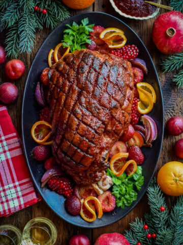 This Festive ham is crosshatched and is plum glazed. It's on platter and is garnished with plums, oranges and festive greenery.