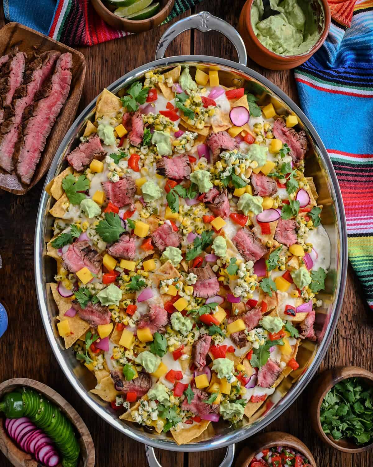An oval platter of Carne Asada Nachos with Elote Salsa & Oaxaca Fondue served with extra toppings.
