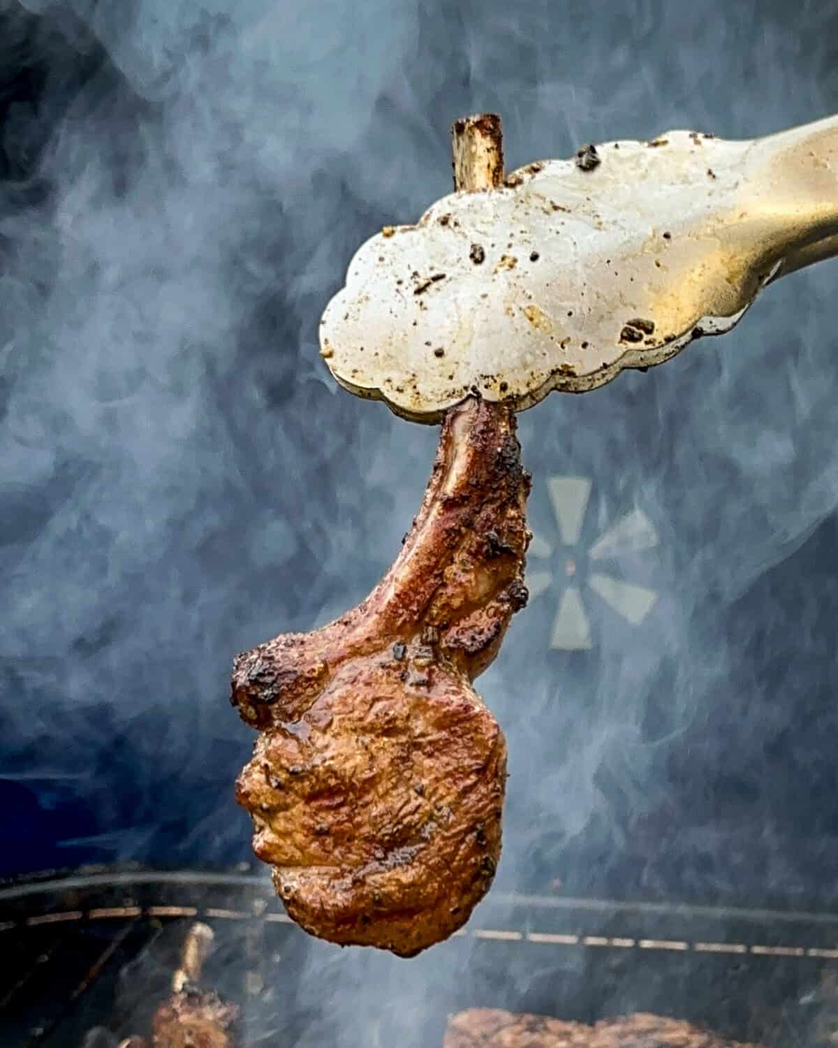 A single lamb lollipop chop is being lifted off a smoky grill.