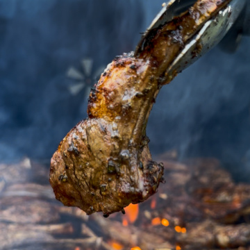 A single lamb lollipop chop is being lifted off a smoky grill.