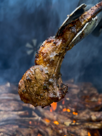 A single lamb lollipop chop is being lifted off a smoky grill.