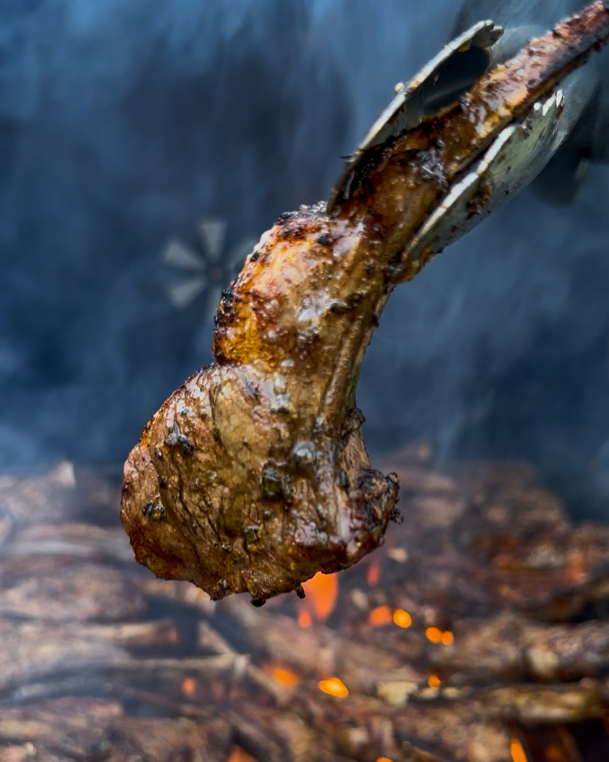 A single lamb lollipop chop is being lifted off a smoky grill.